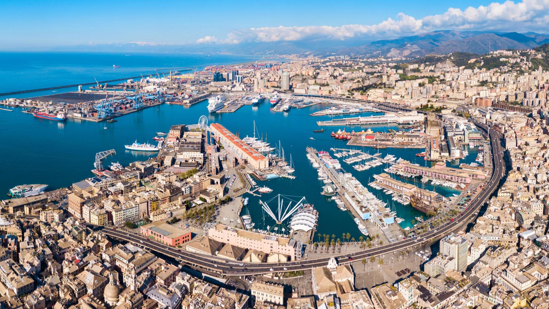 Aerial view of Genoa's port with buildings, ships, and mountains in the background.