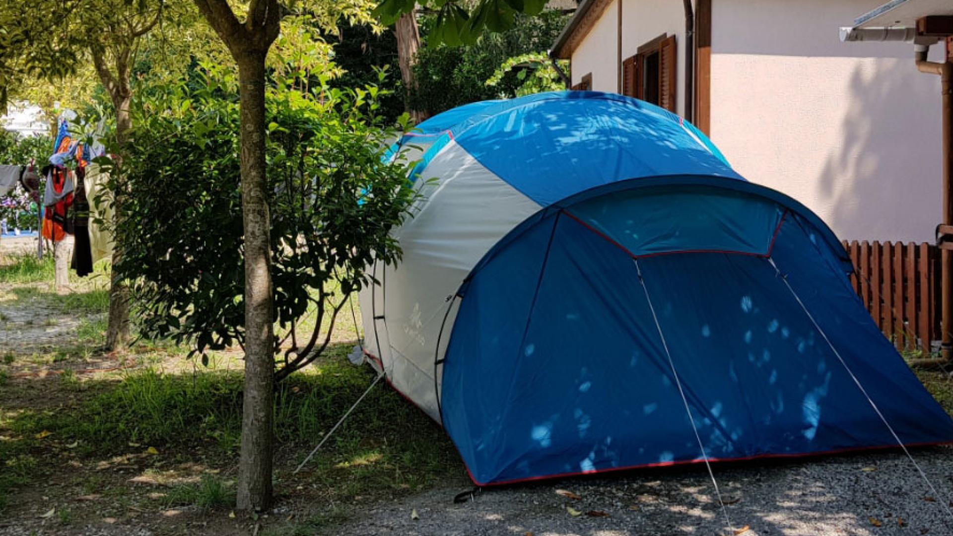 Blue tent at a campsite near a house, surrounded by trees.