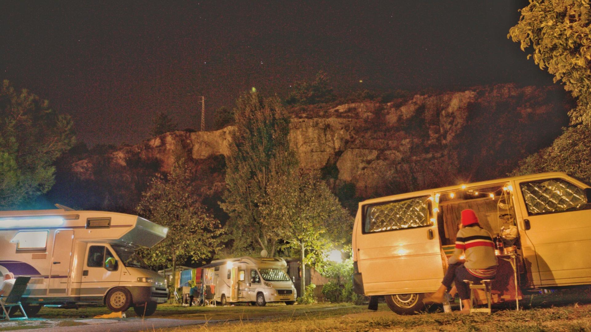 Campeggio notturno con camper e persona seduta vicino a un furgone illuminato.