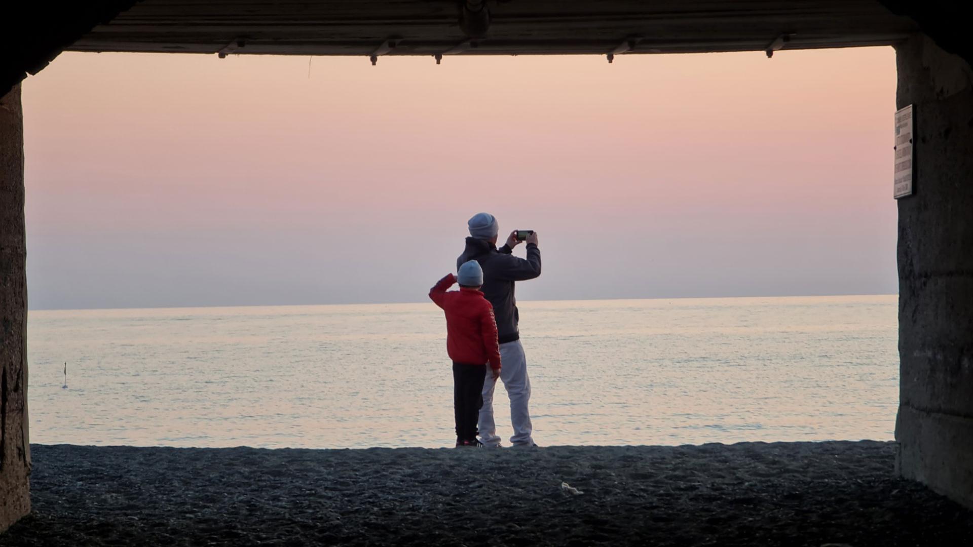Two people admire the sunset on the beach, taking a photo.