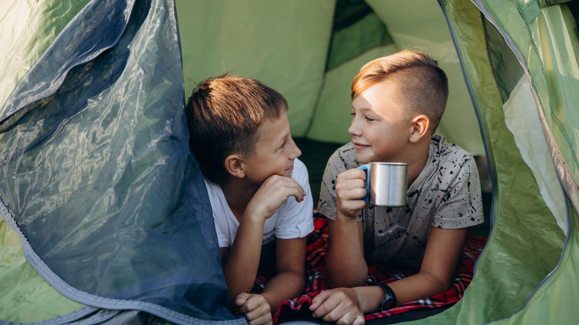 Two kids in a tent, one drinking from a cup.