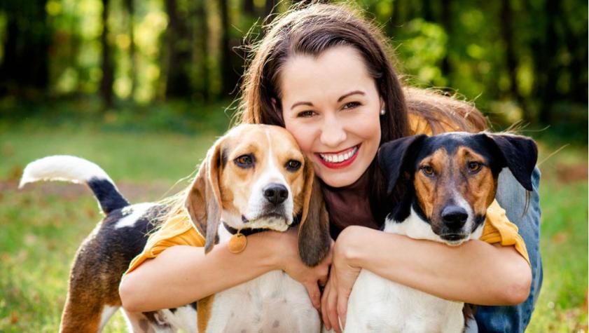 Donna sorridente abbraccia due cani in un parco verde.
