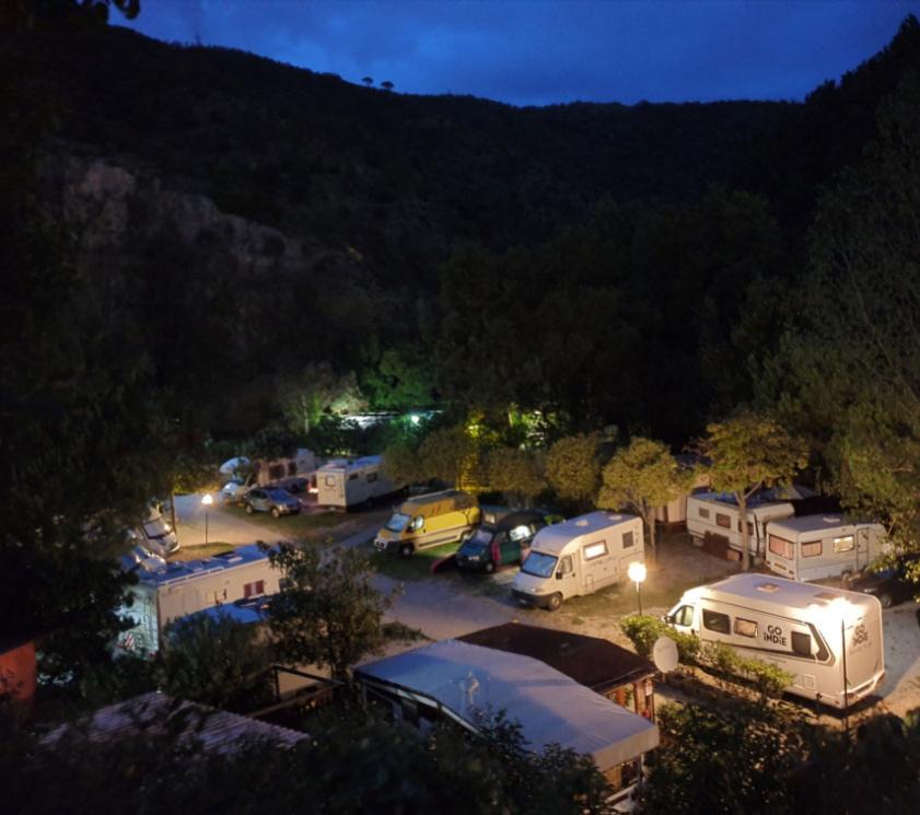 Campeggio notturno con camper illuminati, circondato da alberi e montagne.